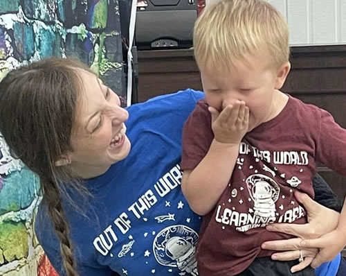 A teacher grinning at the atoshished expression on the face of student seeing lava pour out during a volcano experiment.